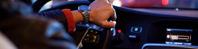A person wearing a wristwatch is driving a car at night. Their hand is on the steering wheel, and the dash displays illuminated controls. The background shows blurred lights from other vehicles, capturing the essence of nighttime city driving—a scene often navigated by personal injury attorneys after incidents.