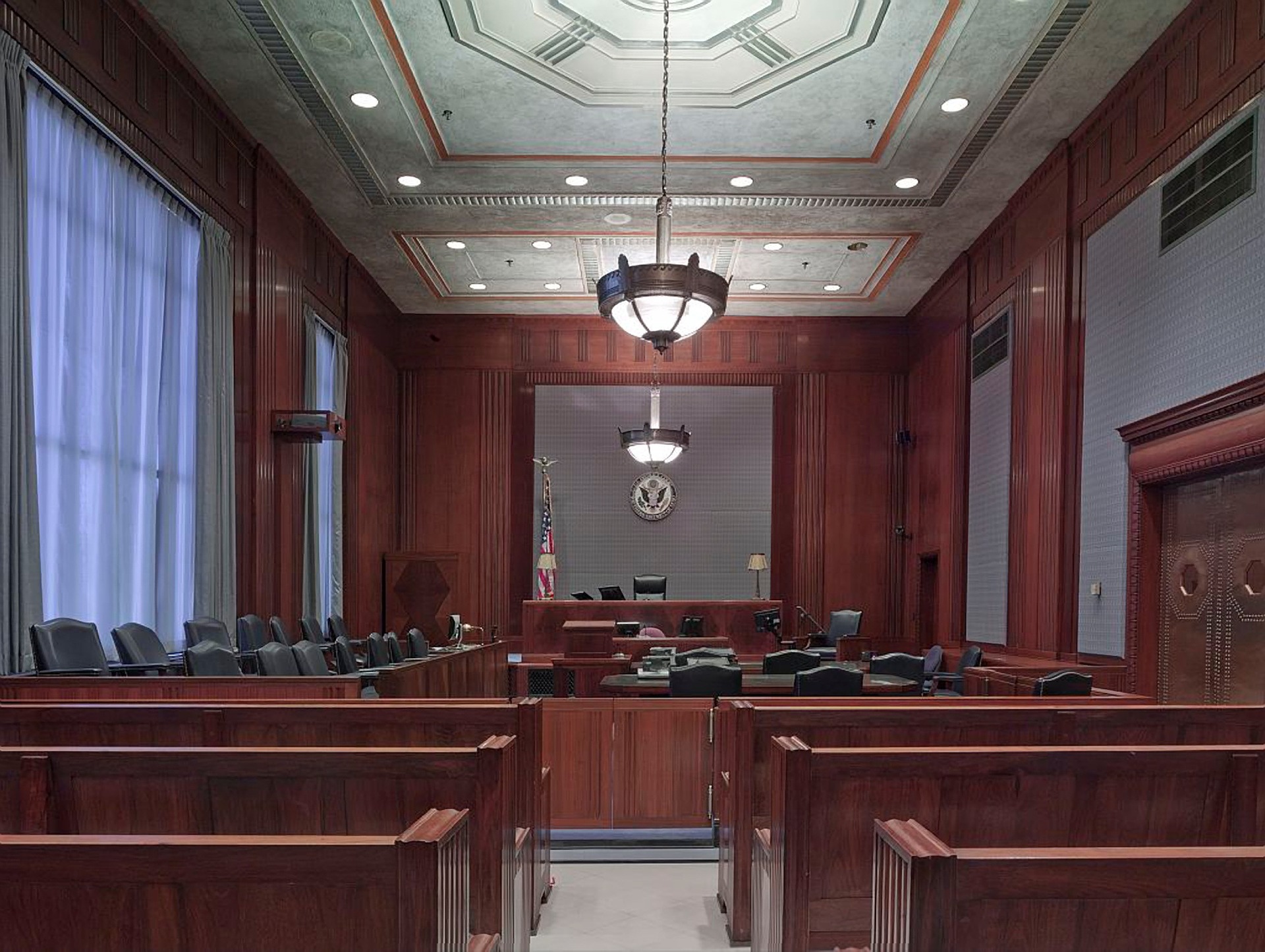 A large, empty courtroom featuring wooden benches, a judge's bench, witness stand, and jury box. The room is lit by overhead chandeliers and natural light from tall windows with blue curtains. The U.S. flag and a seal are displayed behind the judge's bench, ready for cases involving personal injury attorneys.