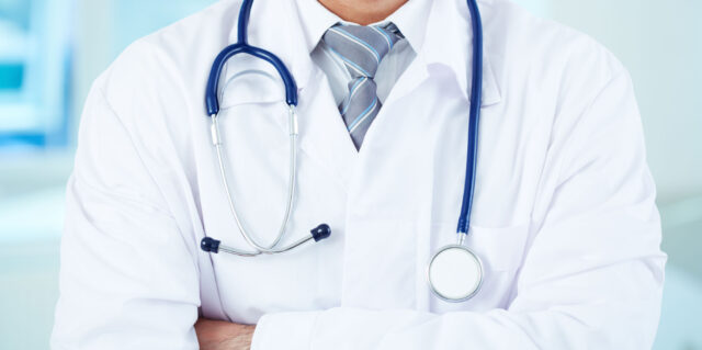 A healthcare professional wearing a white lab coat and a striped tie, with a stethoscope draped around their neck, stands with arms crossed. The background is blurred, suggesting a clinical or medical setting where personal injury attorneys might collaborate to provide comprehensive care.