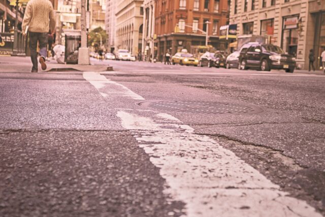 A city street view with a focus on a cracked asphalt road and a manhole cover. A person walks on the left sidewalk, cars are driving and parked along the right side, and buildings line both sides of the street. The overall tone is warm and slightly vintage.