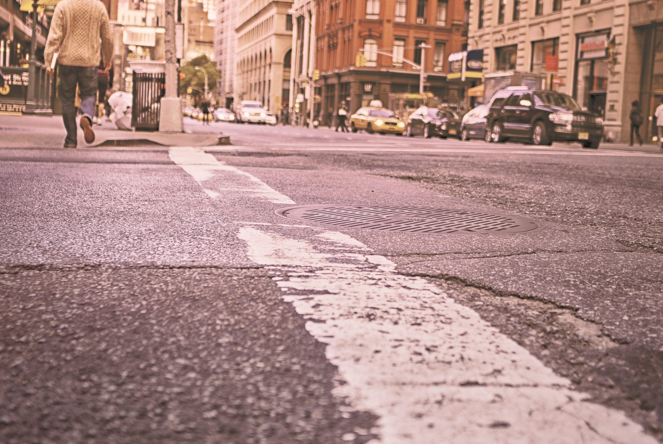 A city street view with a focus on a cracked asphalt road and a manhole cover. A person walks on the left sidewalk, cars are driving and parked along the right side, and buildings line both sides of the street. The overall tone is warm and slightly vintage.