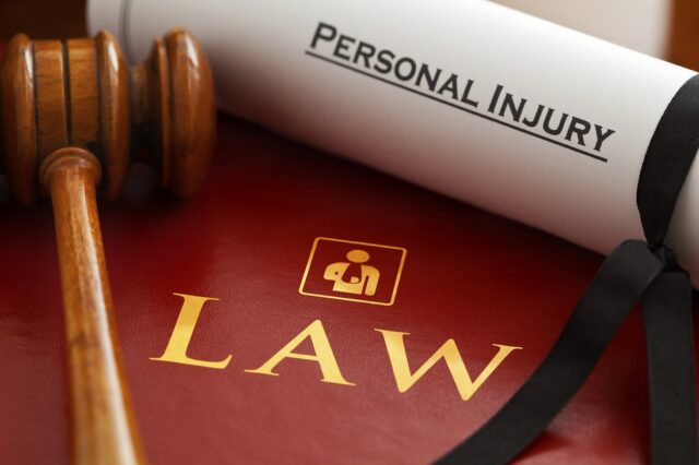 Image of a gavel resting on a red book with the word "LAW" and a briefcase symbol, alongside a rolled-up document tied with a black ribbon labeled "Personal Injury." The items signify legal practices and personal injury claims.