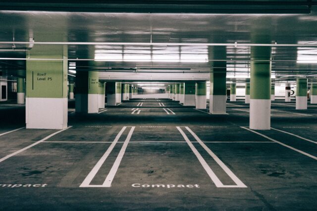 An empty, well-lit underground parking garage with rows of defined parking spaces marked "Compact." Green pillars are interspersed throughout, and signage on the walls indicates "Roof Level P5." The garage appears clean and orderly.