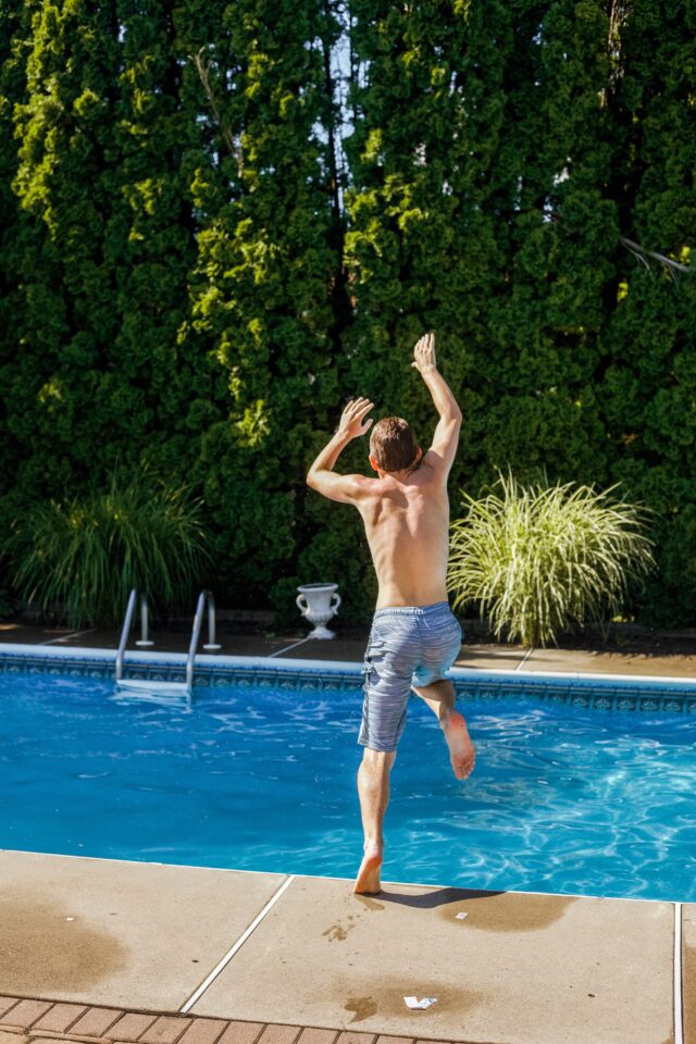 A person with short hair wearing blue swim trunks jumps into a backyard swimming pool next to a ladder. The pool is surrounded by a concrete deck, and there are tall, green bushes and plants lining the fence in the background.
