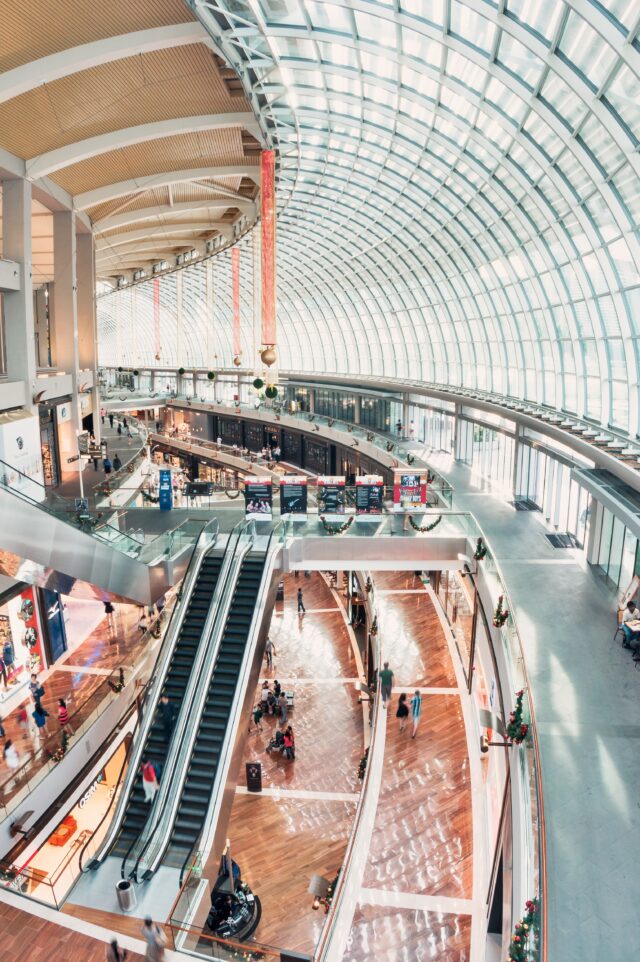 A spacious, modern shopping mall with a glass-domed roof. The mall has multiple levels connected by escalators and features various stores along the walkways. Shoppers are visible on different floors, exploring the shops under the bright natural light.