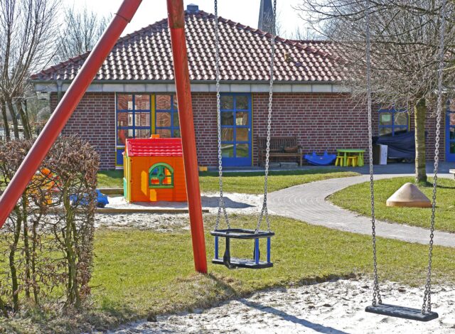 A playground with two empty swings in the foreground. Behind the swings, there is a small, colorful playhouse, and a red brick building with a tiled roof. Trees without leaves surround the area, and a paved path winds through the grass.