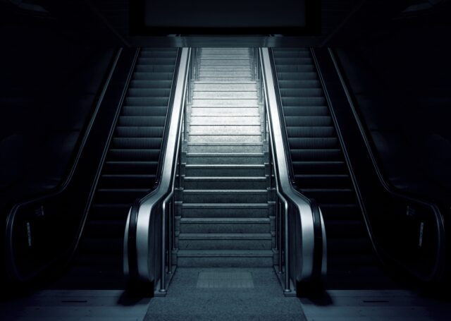 A dimly lit indoor space shows a symmetrical view of two escalators flanking a central staircase. The escalators are not in motion, and the scene has a moody, almost futuristic feel with a strong emphasis on shadows and light.