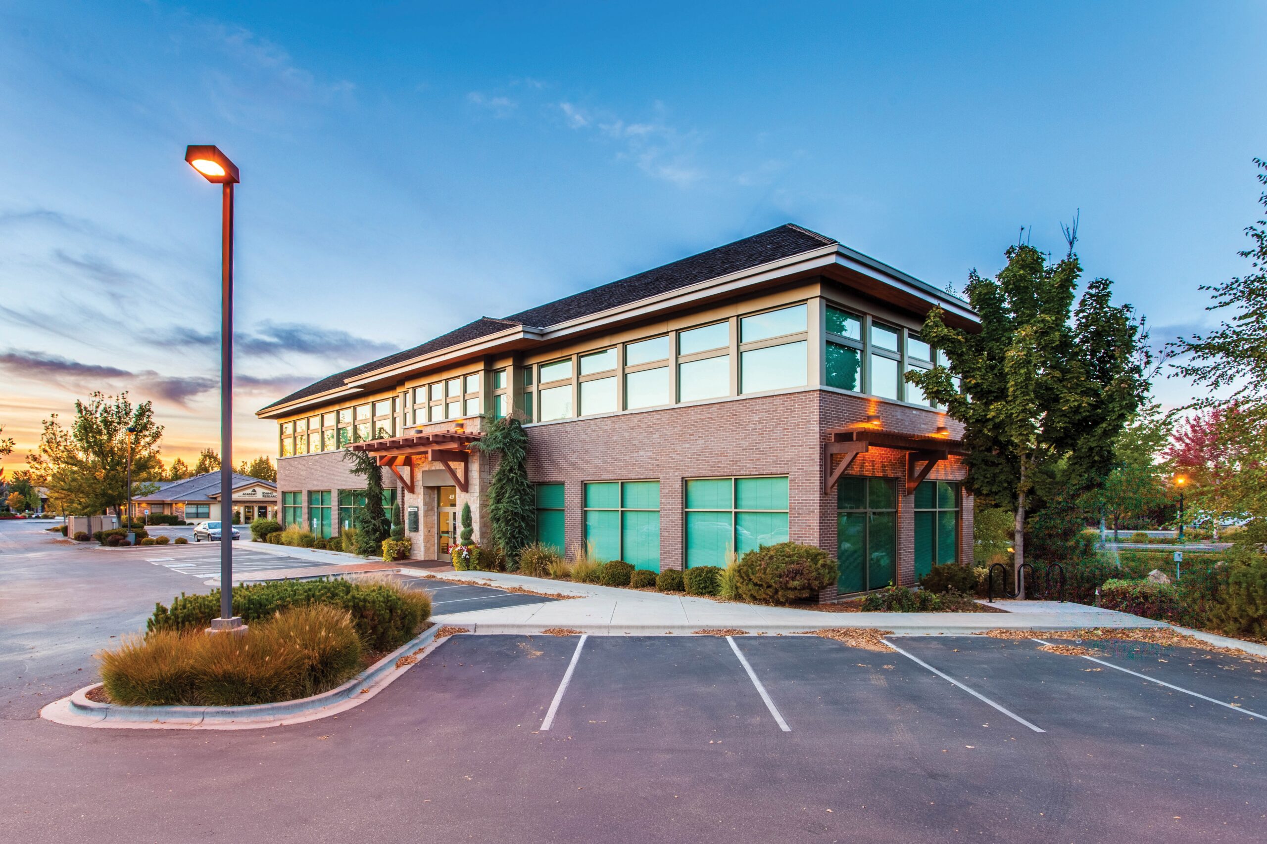A modern, two-story building with large windows and a brick facade, surrounded by greenery. A well-lit parking lot with a few cars is in front. The sky is partly cloudy during a sunset, casting a warm glow on the scene.