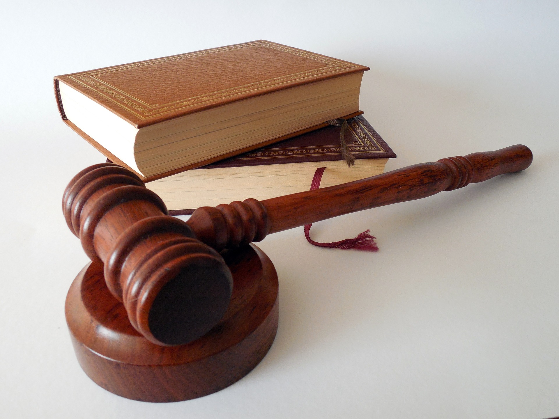 A wooden gavel rests on its sounding block in the foreground, with a stack of closed books, likely legal texts, in the background. The books have leather covers and one has a bookmark peeking out. The scene is set on a clean, white surface.