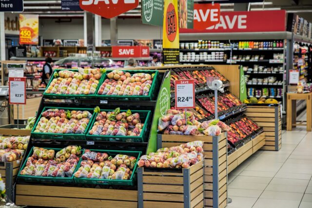 A supermarket produce section with multiple displays of prepackaged fruits, mainly apples, marked with price tags. Shelves in the background have various canned and packaged goods. Red "SAVE" signs hang above, indicating promotions or discounts.