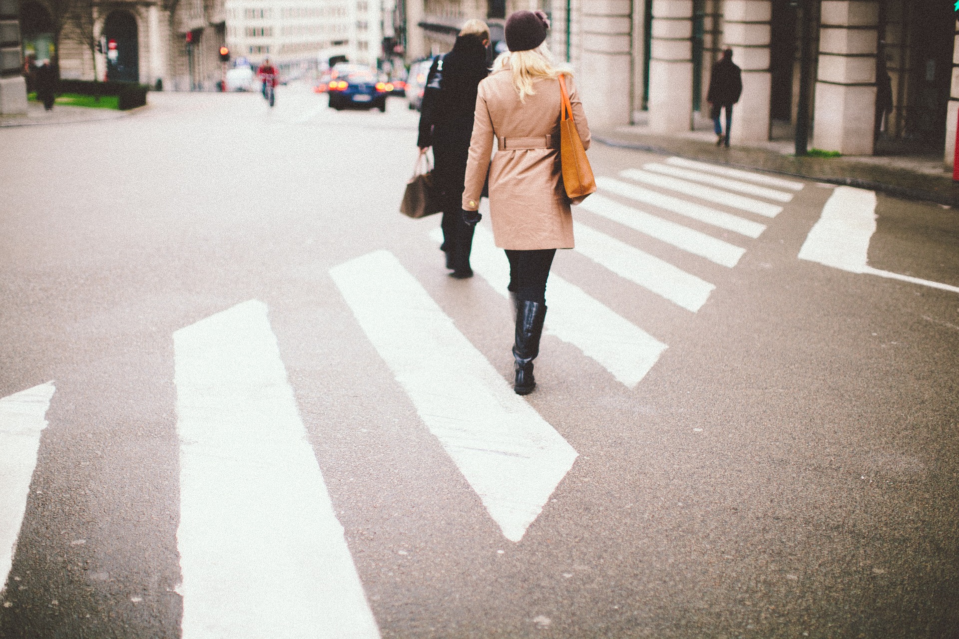 A person wearing a beige coat, black boots, and a black hat walks along a zebra crossing on a city street. Other people and buildings are visible in the background.