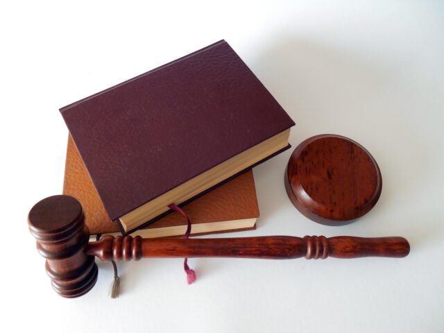 A wooden gavel, a sound block, and two closed books with brown and burgundy covers lie on a white surface. A red ribbon bookmark is visible between the pages of one book. The gavel and sound block appear polished and are placed near the books.