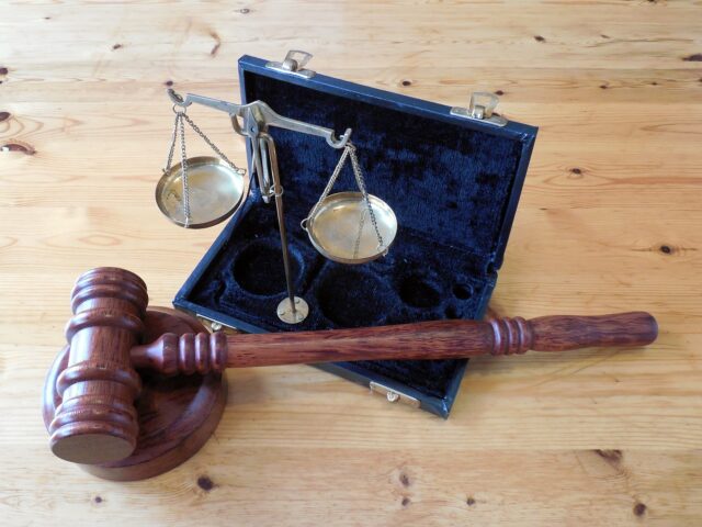 A wooden gavel with a round base is placed in front of an open black case containing a brass balance scale. The background is a wooden surface. The gavel and scale symbolize justice and law.
