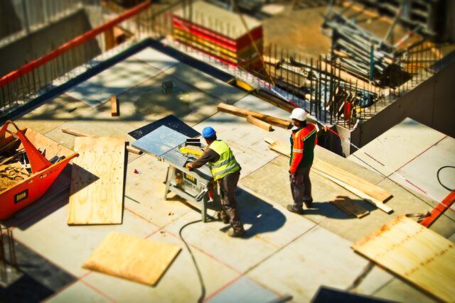 Two construction workers on a building site with scattered wooden planks and materials. One worker uses a table saw while the other stands nearby. Both are wearing safety helmets and protective vests. The environment has safety barriers and equipment visible.