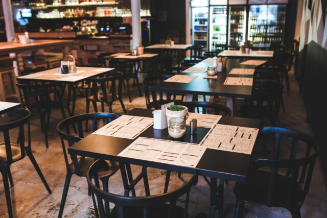 A modern restaurant with black tables and chairs, set with menus, napkins, and small potted plants in the center. The bar area is in the background, along with refrigerators stocked with drinks. The ambiance is cozy with wooden and industrial elements.