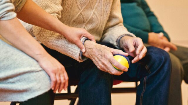 An elderly person in a knitted sweater holds a small yellow and blue ball, while another person in a gray sweater gently touches their arm, offering support. A third person in a green sweater is seated beside them, slightly out of focus.