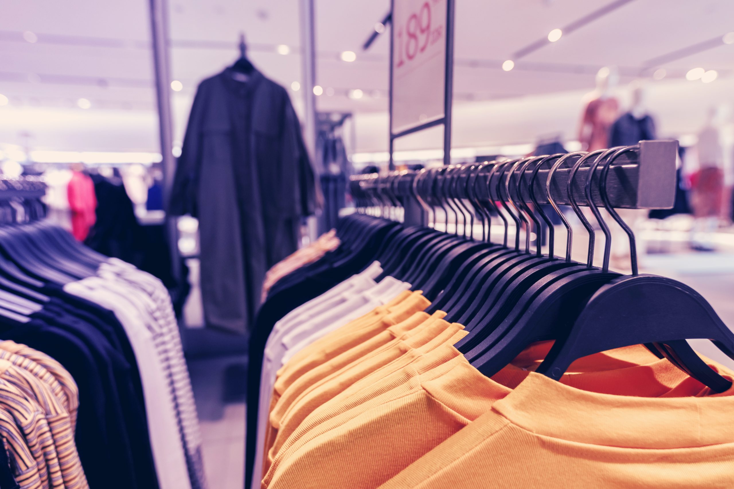 A clothing store display showing various garments on hangers. The focus is on a row of brightly colored tops, predominantly yellow, with more clothing items and racks visible in the background within a modern, well-lit store setting.
