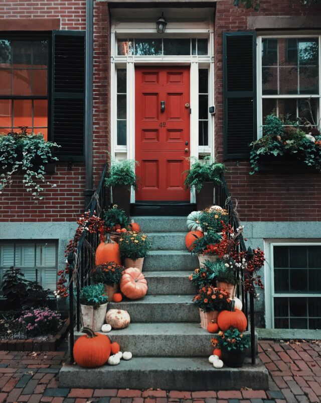 A brick house with a red door decorated for autumn. The steps leading to the door are adorned with various pumpkins, gourds, chrysanthemums, and autumn foliage. Greenery and flower pots are arranged on both sides of the steps and windows.