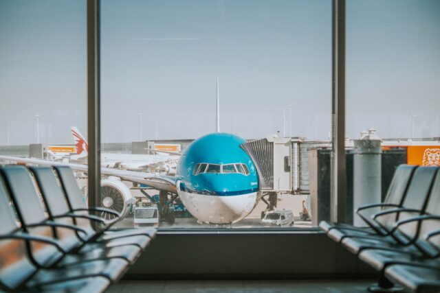A blue and white airplane is parked at a gate with a jet bridge attached at an airport terminal. The view is seen through large glass windows flanked by rows of empty seats, with other airport facilities and planes visible in the background.