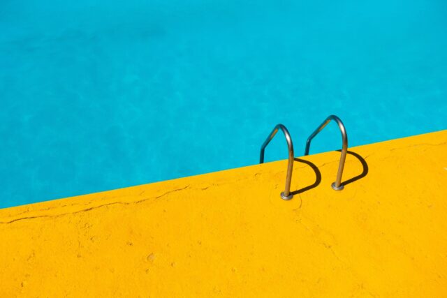 A vibrant, bright yellow pool deck contrasts with the clear blue water of a swimming pool. A metal ladder descends into the pool, creating a simple yet striking geometric composition.