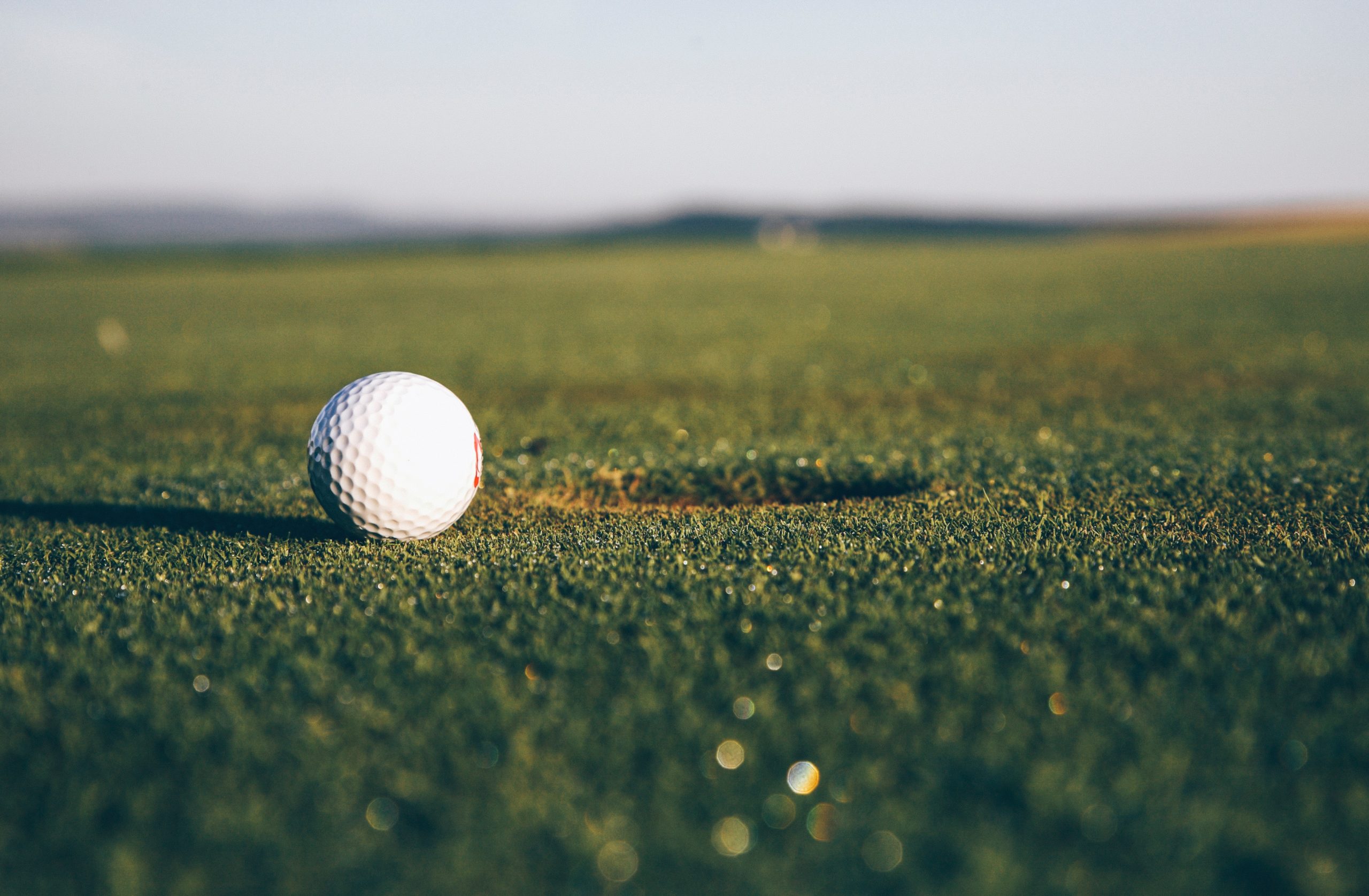 Close-up of a white golf ball resting on the green grass near a hole on a golf course, with a blurred background. The photo is taken at a low angle, highlighting the ball's details and the texture of the grass.
