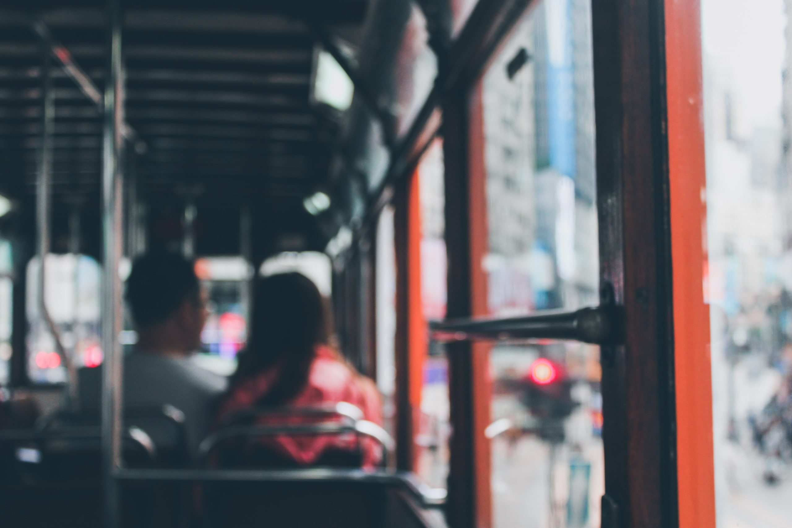 Two people sit next to each other on a bus or tram with an orange and wooden interior. The background features the blurred, bustling cityscape seen through the open window, enhancing the sense of movement and urban life. Both individuals are slightly out of focus.