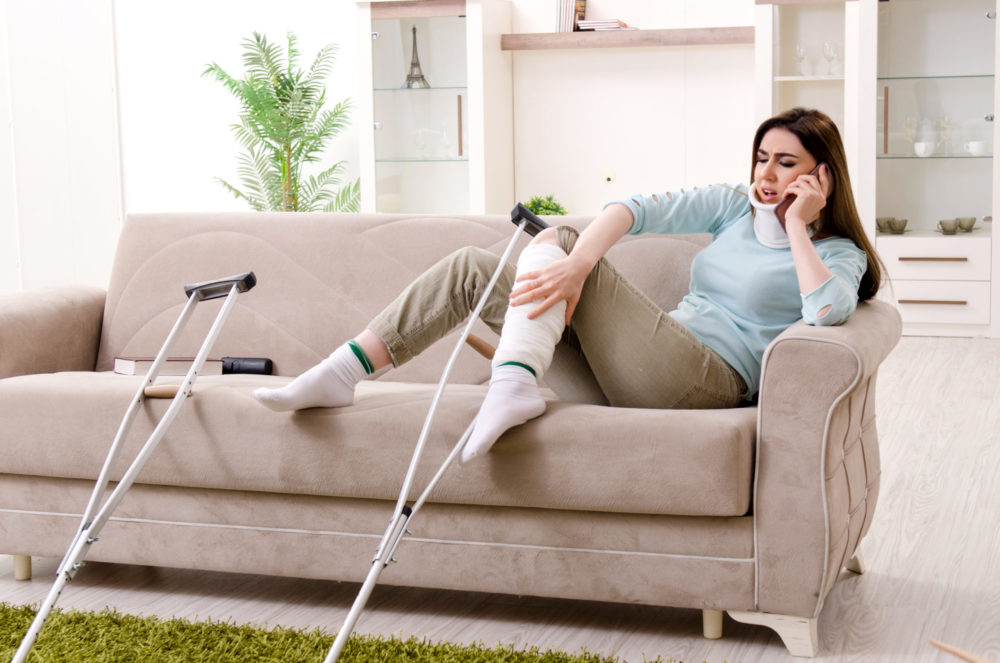 A woman with a bandaged leg sits on a beige couch, holding a phone to her ear. She appears to be in conversation. Two crutches lean against the couch, and a small table with books is beside her. The room is well-lit with a potted plant in the background.