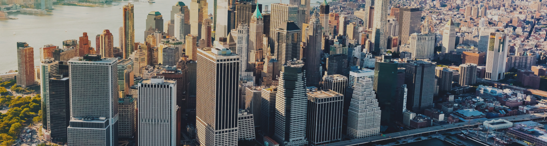 An aerial view of a densely packed cityscape featuring numerous soaring skyscrapers and tall buildings. The scene includes a mix of urban architecture with a river visible in the background and green park areas scattered among the buildings, hinting at the bustling life where vehicle accident attorneys may find their clients.