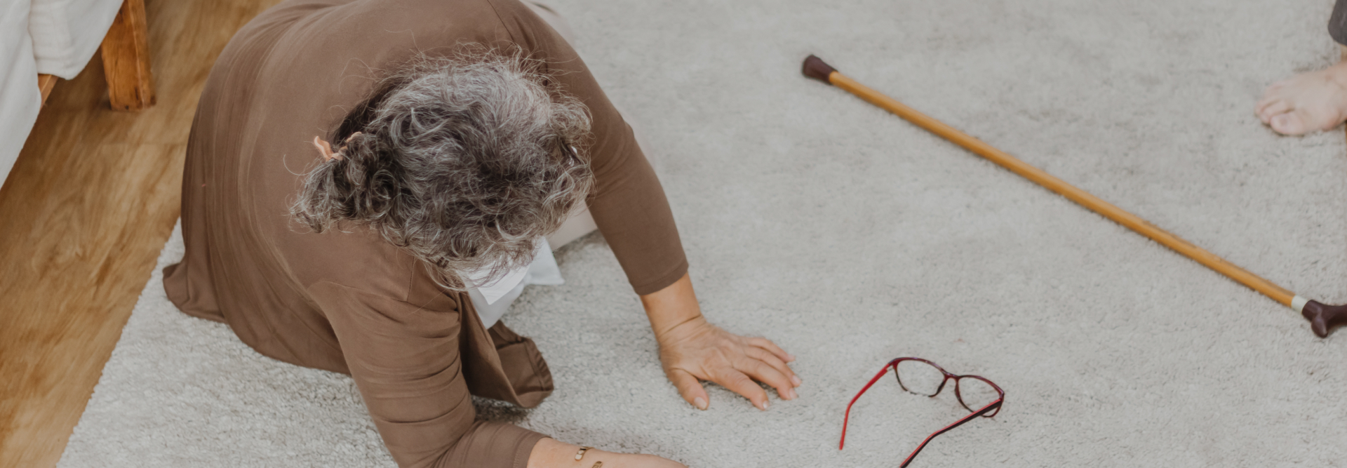 An elderly person with gray hair, wearing a brown sweater, is lying on the floor next to a walking cane and red-framed glasses. The scene suggests the person may have fallen. Personal injury attorneys often handle cases like these to ensure justice and compensation for those affected.