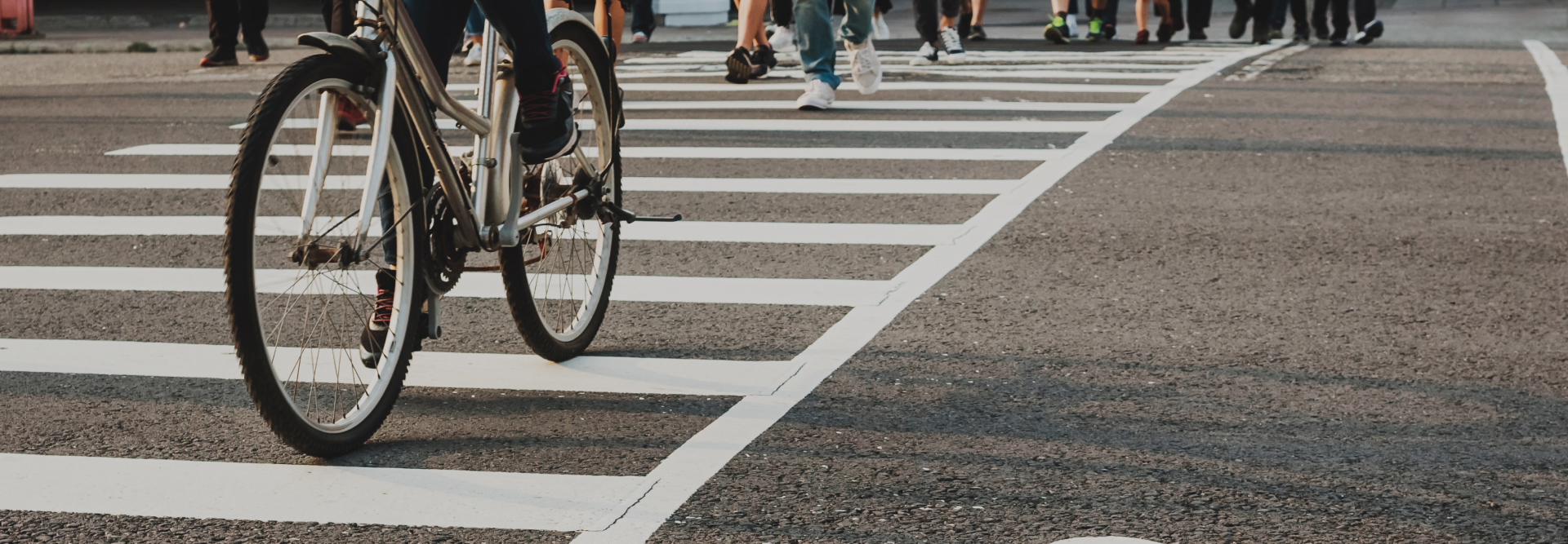 A close-up of a bicycle and a person riding it on a crosswalk, alongside several pedestrians crossing the street. The image captures the lower halves of the individuals, focusing on their legs and footwear. The scene appears to be in an urban setting where personal injury attorneys are often consulted for accidents.

