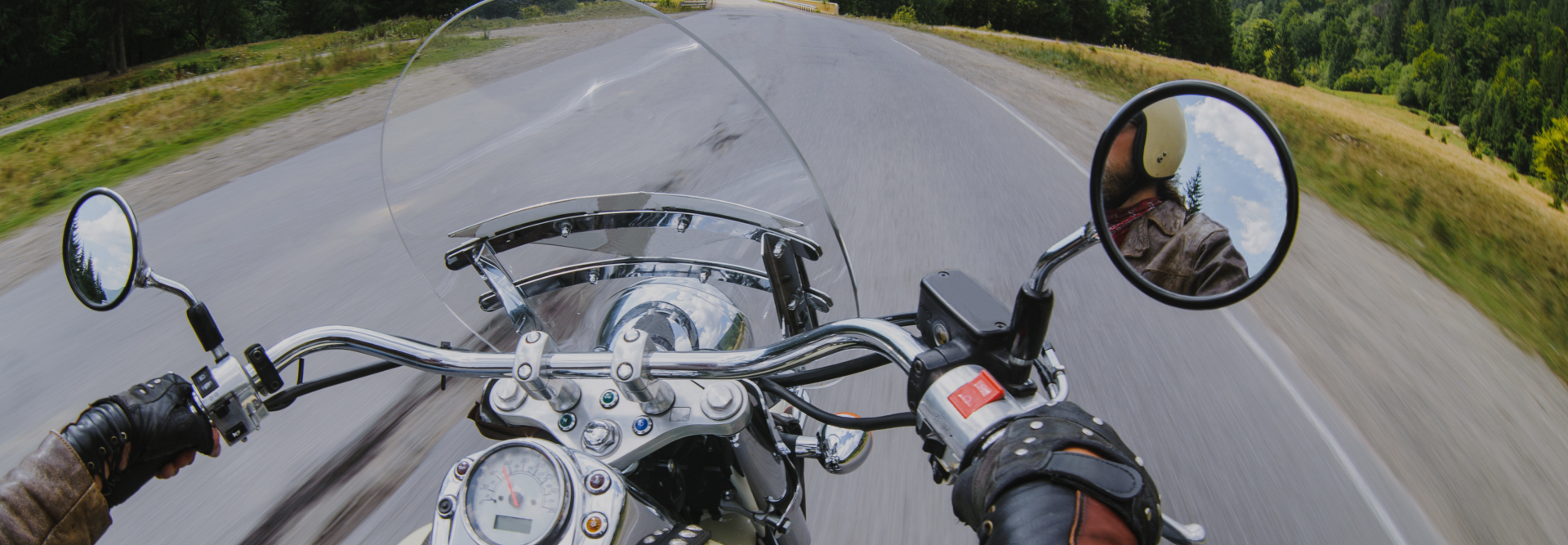 A motorcyclist is riding on an open road with trees and greenery on the side. The shot captures the rider's perspective, showing the handlebars, speedometer, rearview mirrors, and the road ahead. The landscape is blurred, indicating motion; Westchester County Motorcycle Accident Attorneys concept image