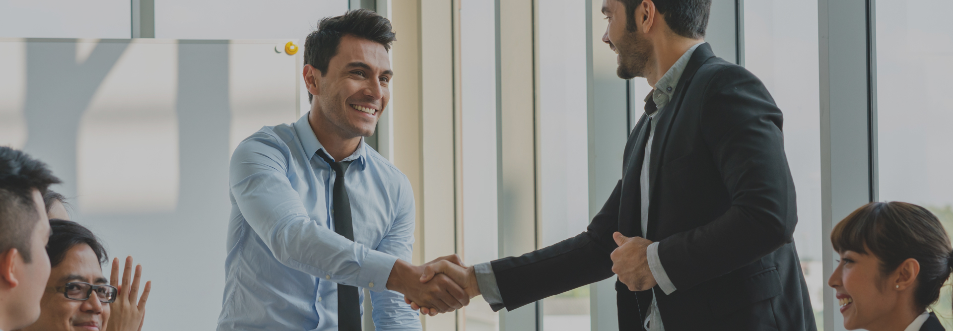 Two men are shaking hands in a well-lit office meeting room, surrounded by colleagues who are smiling and clapping. One man is wearing a blue shirt and tie, the other a dark suit. This scene suggests a successful business agreement between prominent vehicle accident attorneys or the conclusion of a positive meeting.