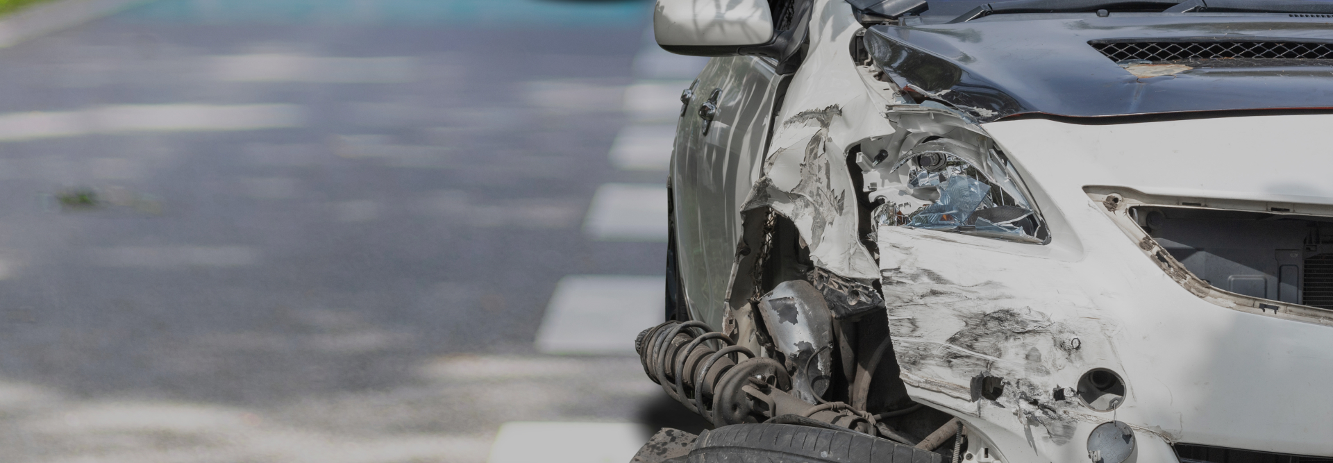 A close-up of a damaged white car's front right corner, showing smashed headlight and crumpled metal. The car seems to have been in a collision and is partially on a road after a collision with an MTA bus; Westchester County Auto Accident Attorney concept image