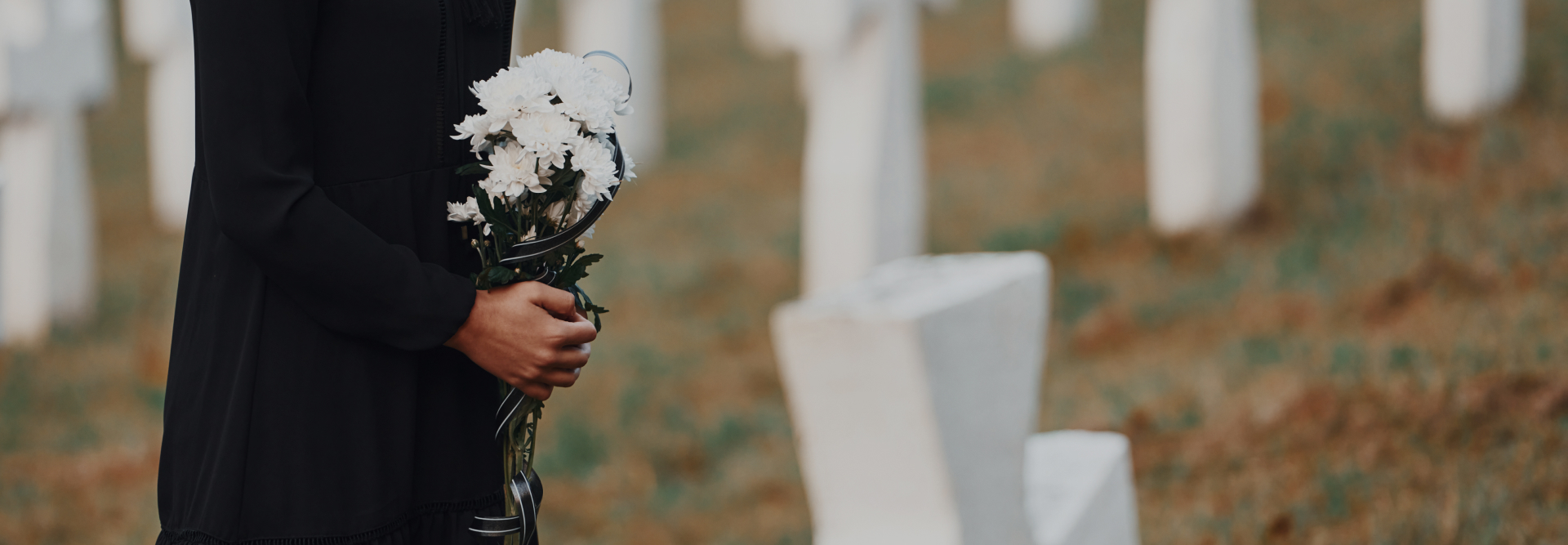 A person dressed in black holds a bouquet of white flowers while standing in front of a grave, with more graves visible in the background. The setting appears to be a cemetery, possibly mourning a loved one lost in an accident. Nearby, injury lawyers could be seen assisting other grieving families.