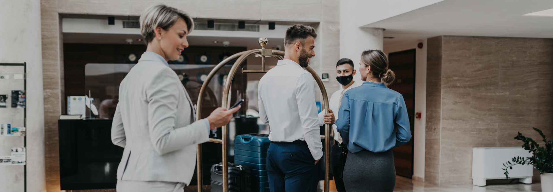 A smiling woman checks her phone in the foreground while two men in suits and a woman in a blue blouse talk near a luggage cart in what appears to be a hotel lobby. One man, possibly a vehicle accident attorney, is wearing a face mask. The setting is modern, with neutral tones and contemporary decor.