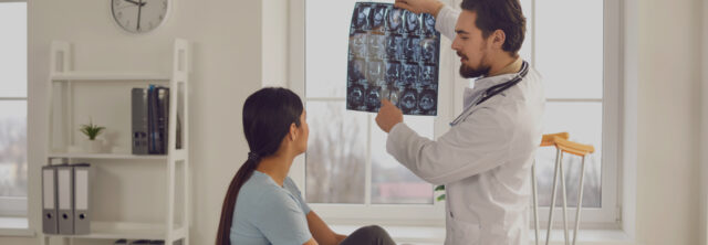 A doctor wearing a white coat and stethoscope shows a patient x-ray images in a medical office. The patient, seated, looks attentively at the x-rays. The room has large windows, a clock, books, and crutches in the background—a scene often referred to by personal injury attorneys when discussing medical consultations post-accident; Port Chester Personal Injury Attorney / Mount Vernon Personal Injury Attorneys concept image