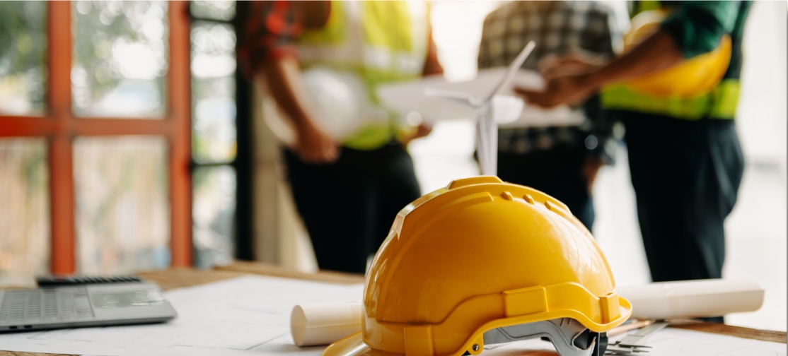 A yellow construction helmet rests on a table with blueprints and a calculator. In the blurred background, three people in safety vests have a discussion, holding a paper and a tablet, suggesting the assistance of personal injury attorneys for construction site planning meetings.