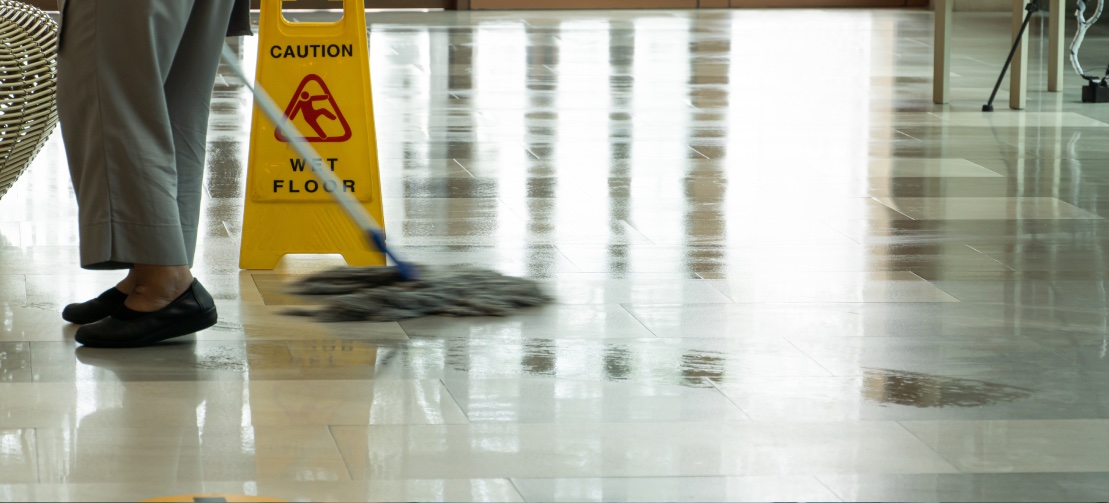 A person in gray pants and black shoes mops a shiny floor next to a yellow 
