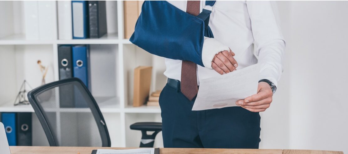 A person wearing a white shirt and maroon tie is holding a document in their right hand, while their left arm is in a blue sling. They are standing near an office desk with shelves containing binders and books, possibly seeking advice from personal injury attorneys.