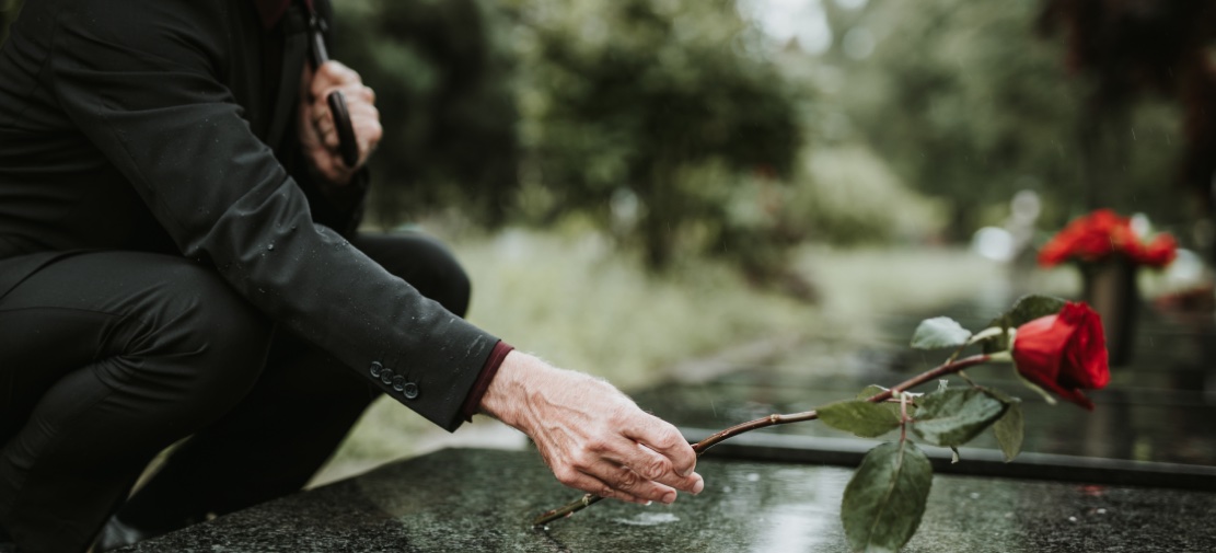 A person dressed in black holds a red rose while kneeling and placing it on a gravestone in a cemetery. The background is blurred with greenery and colorful flowers on other gravestones. The atmosphere appears solemn and reflective, reminding one of the delicate nature of life frequently encountered by personal injury attorneys.