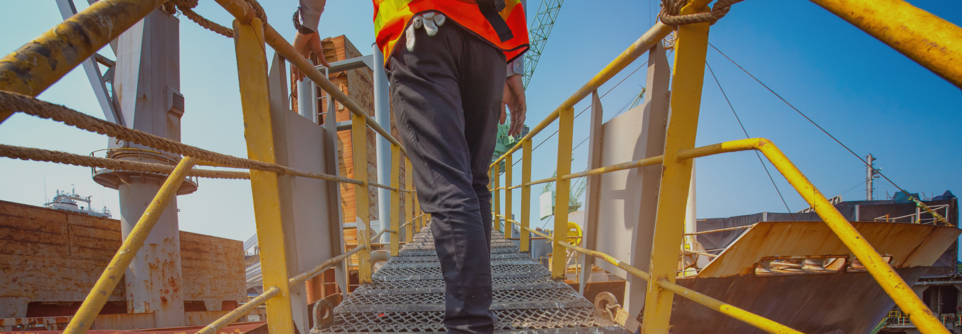 A person wearing a red safety vest and black pants walks up a yellow metal staircase at a construction or industrial site. Machinery and scaffolding are visible in the background under a clear blue sky, reminiscent of scenes often reviewed by vehicle accident attorneys during site inspections.