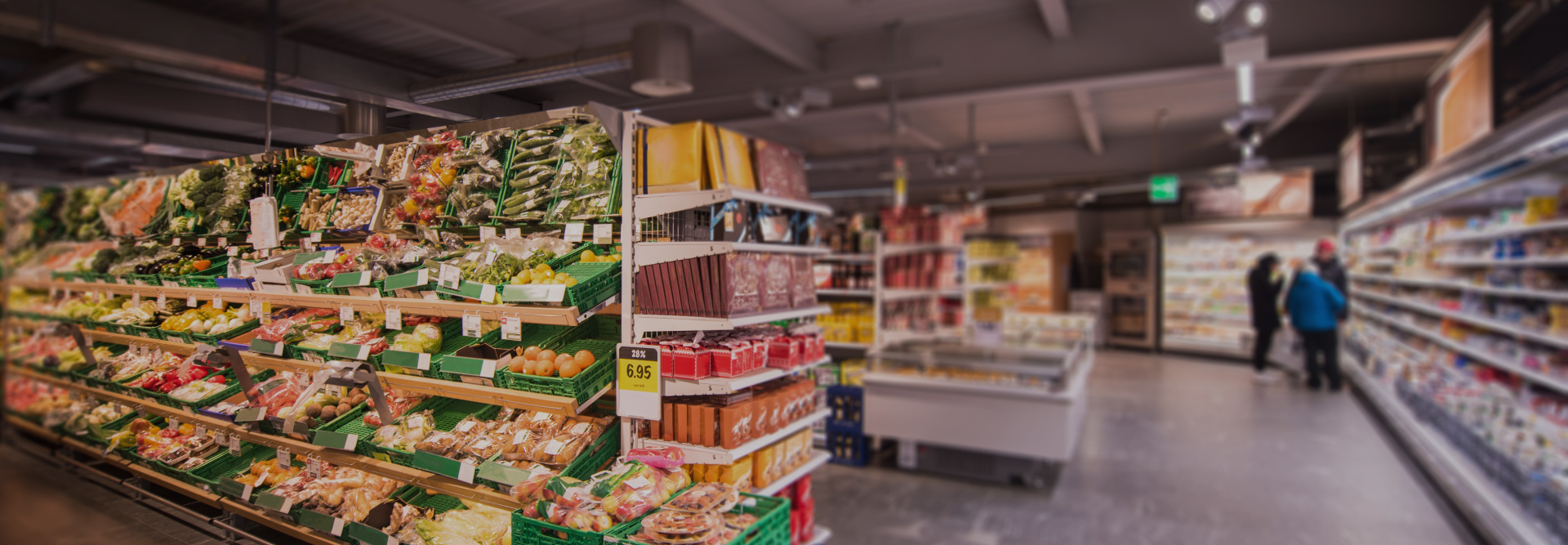 A brightly lit supermarket aisle featuring shelves stocked with various fresh vegetables, fruits, and packaged groceries reflects the orderliness you'd expect from dedicated injury lawyers. Two shoppers are seen in the background near the refrigerated section. The floor is clean, and the shelves are neatly organized.