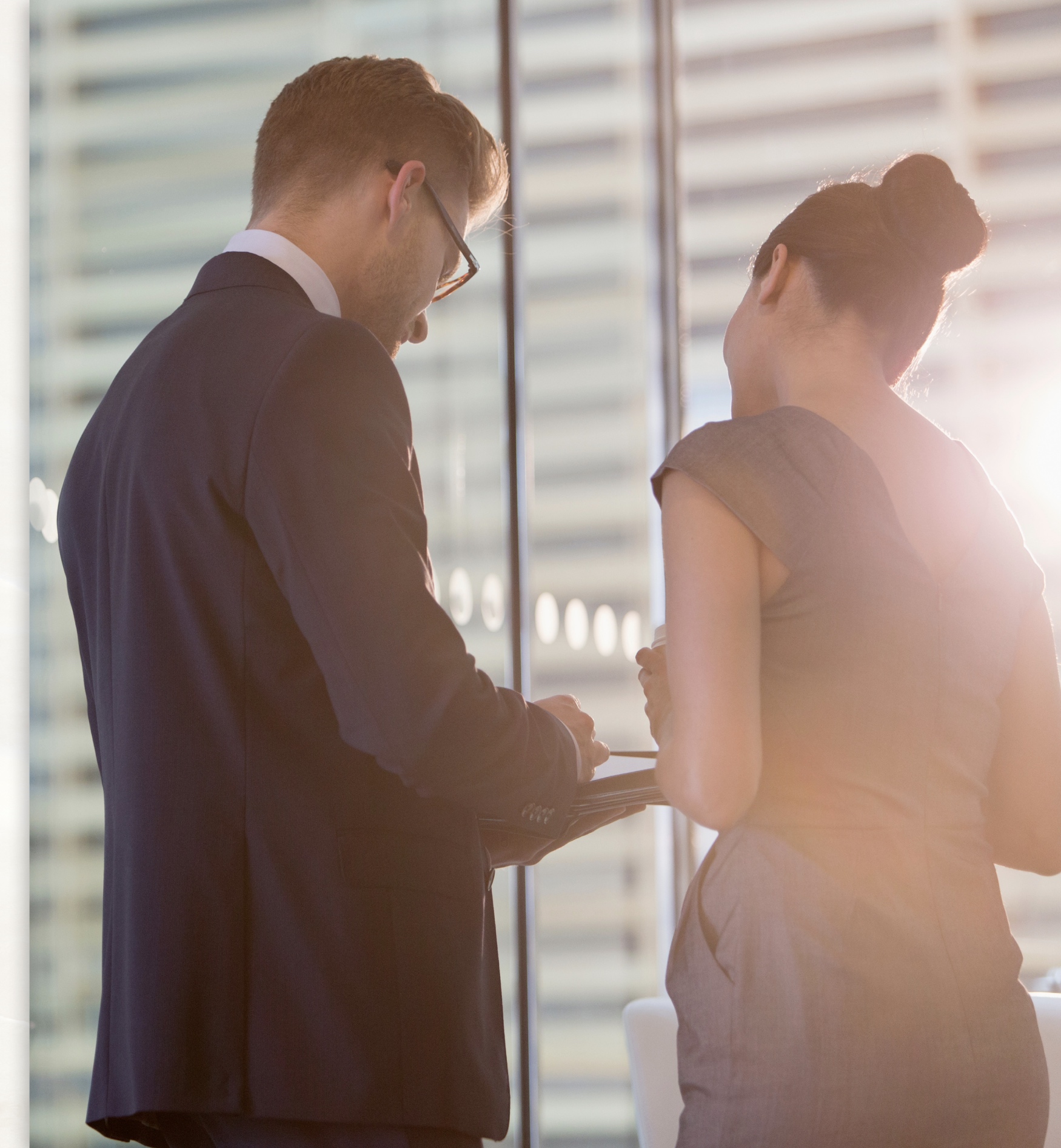 Two professionals in formal business attire, identified as vehicle accident attorneys, stand next to a large window in a modern office. They are engaged in discussion, and one holds a tablet or document. Sunlight streams through the window, creating a bright and airy atmosphere.